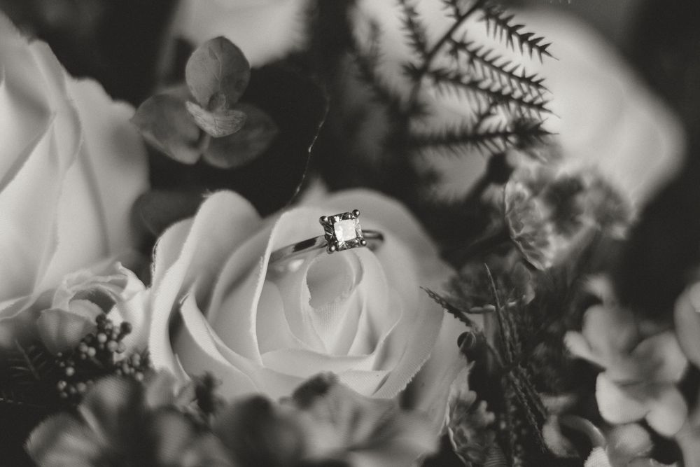 Black and white close-up of princess-cut diamond engagement ring resting on white rose bouquet.