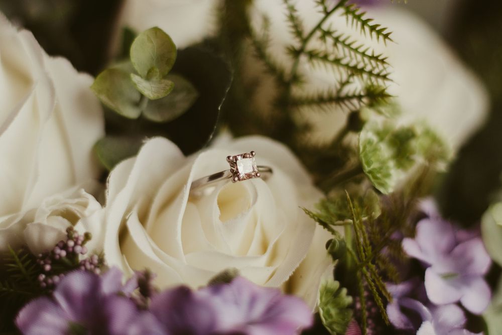 Princess-cut diamond engagement ring nestled in white roses and purple wildflowers bridal bouquet.