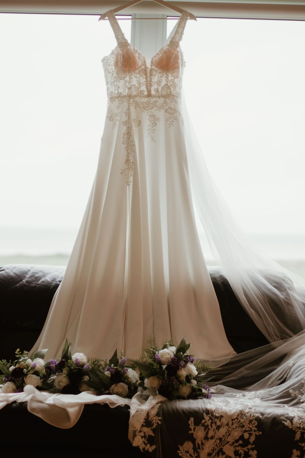 Elegant lace A-line wedding gown hanging by window with floral bouquets and veil below.