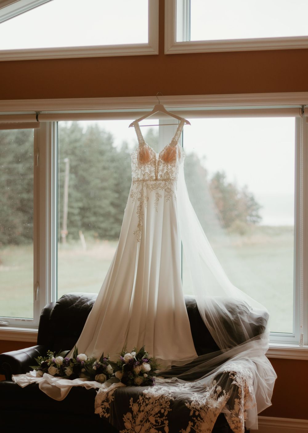 Romantic lace bridal gown hanging in window light with bouquets and veil draped below.