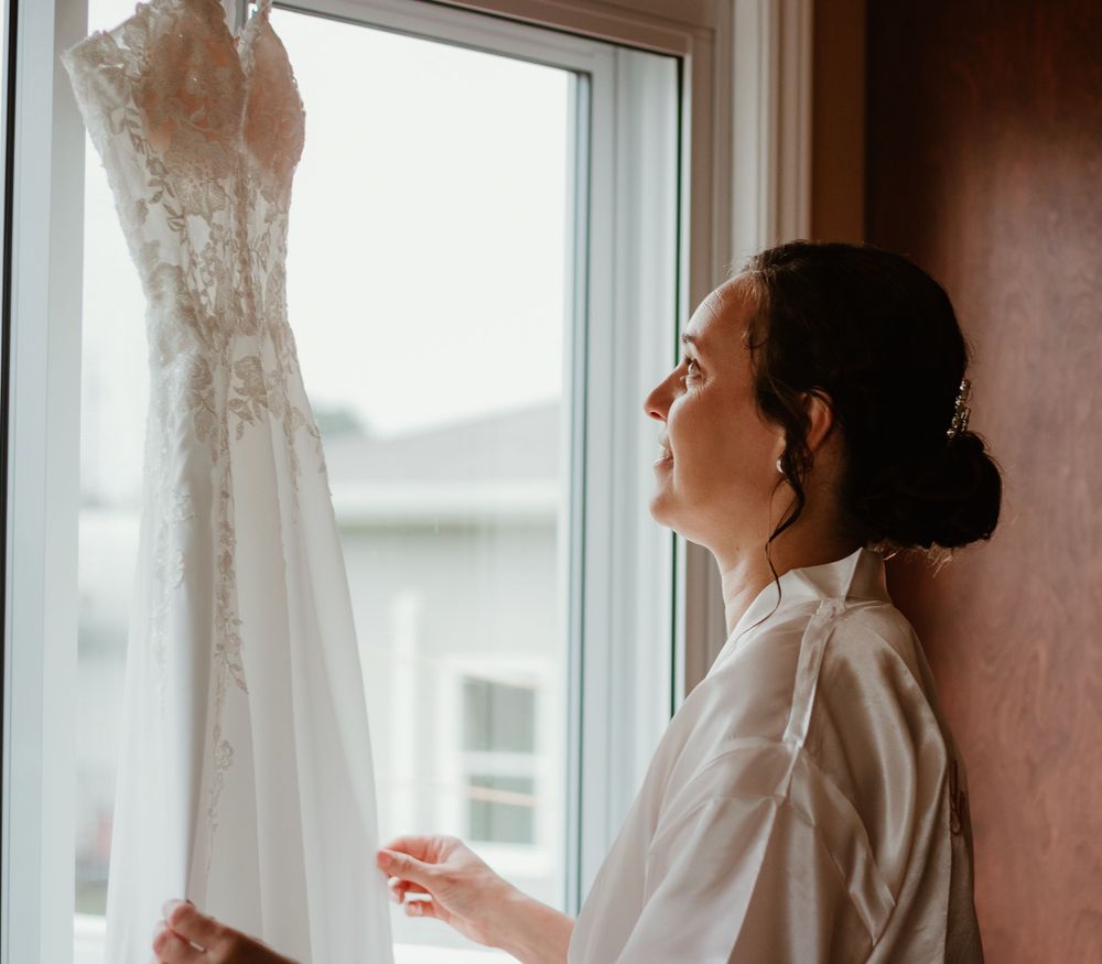 Bride in white robe admiring her lace wedding dress hanging by a window on her wedding day.