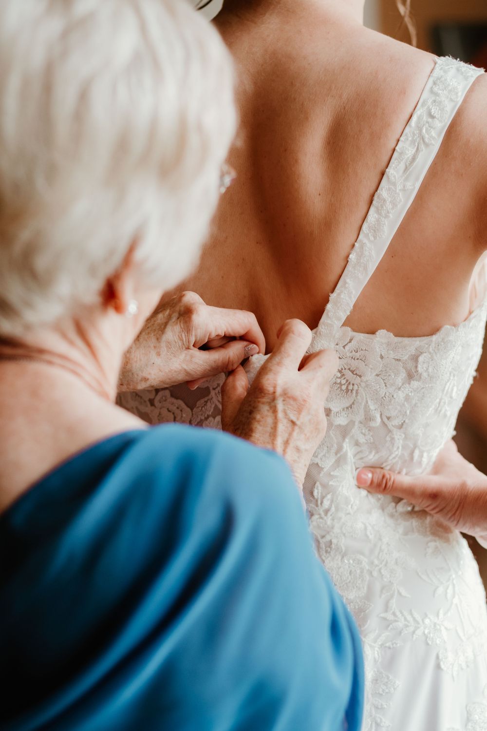 Older woman in blue dress carefully buttoning the back of a bride's lace wedding gown.