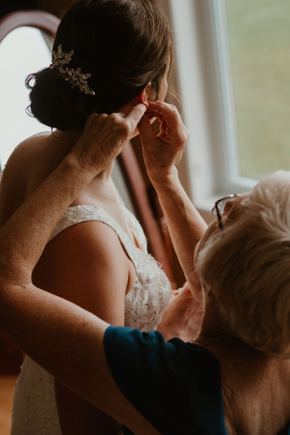 Mother helping bride put on earrings while bride wears lace gown with floral hair accessory.