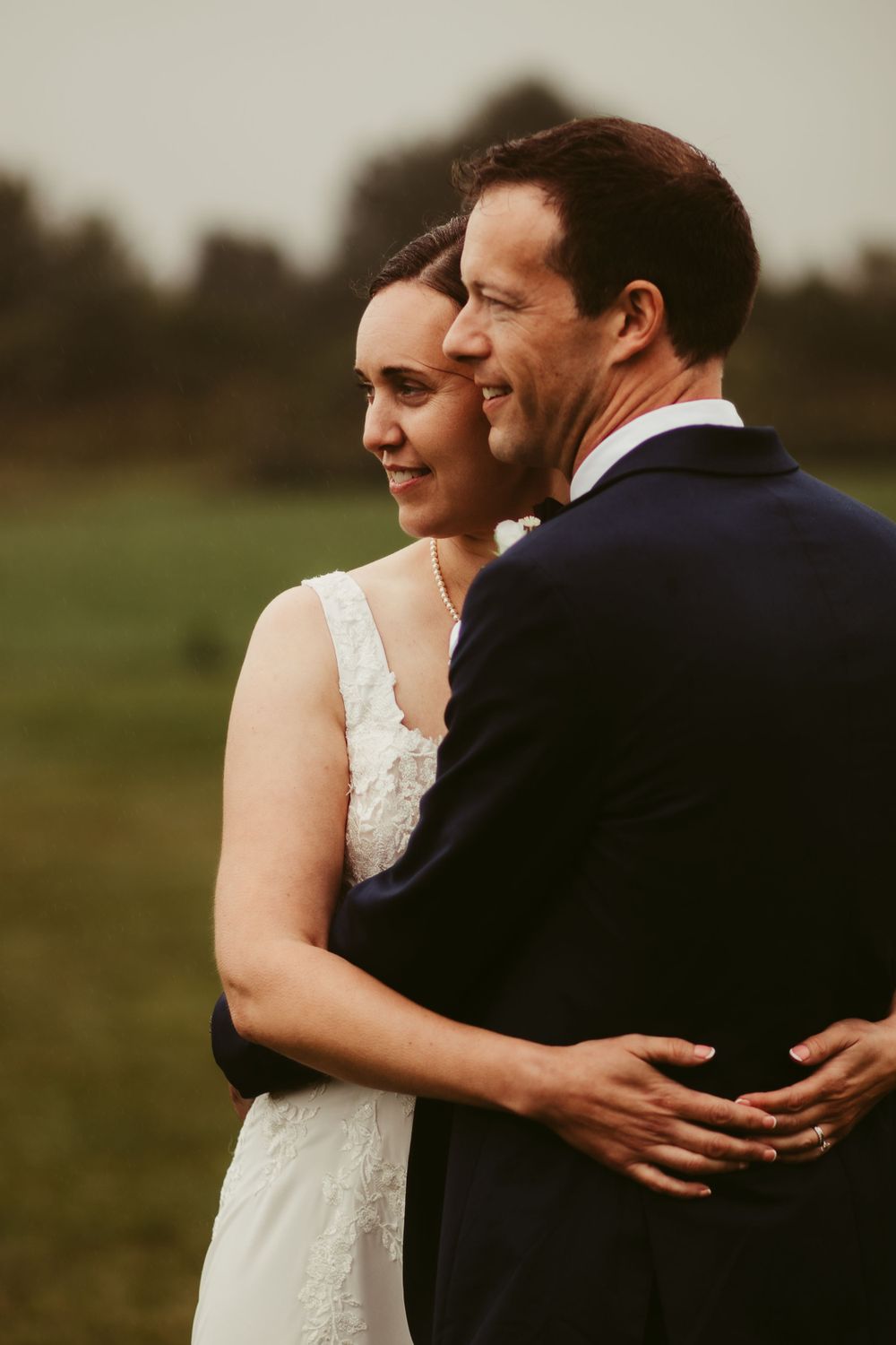 Bride and groom embracing outdoors, with the bride in a lace gown smiling as the groom holds her close.
