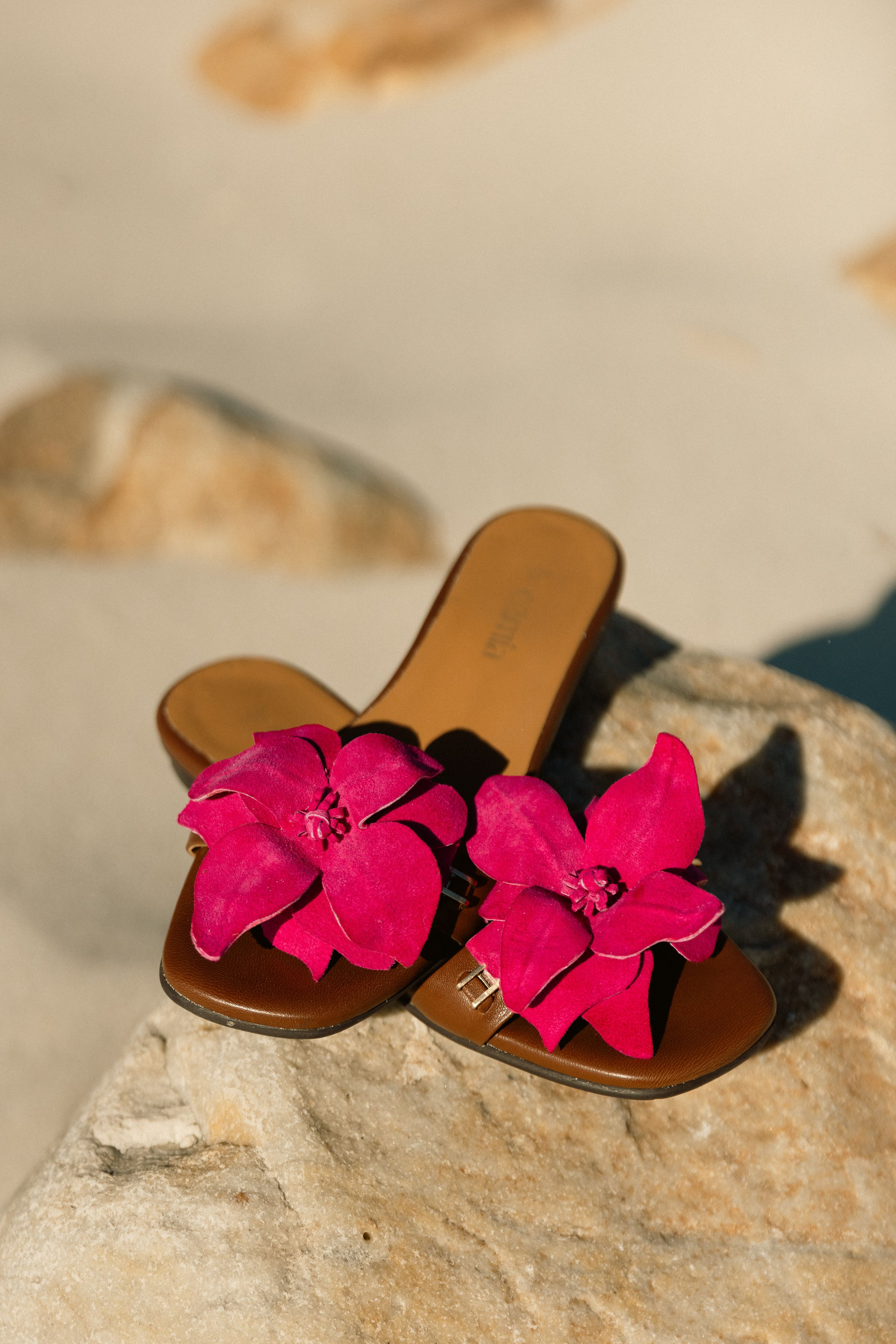 Brown leather sandals with vibrant pink flower decorations displayed on a light stone surface.