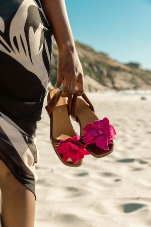 Hand holding brown sandals with pink flowers against a sandy beach backdrop.