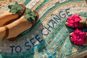 Sandals with green and pink flowers arranged on a blue mosaic tile with text reading 'TO SEE CHANGE'.