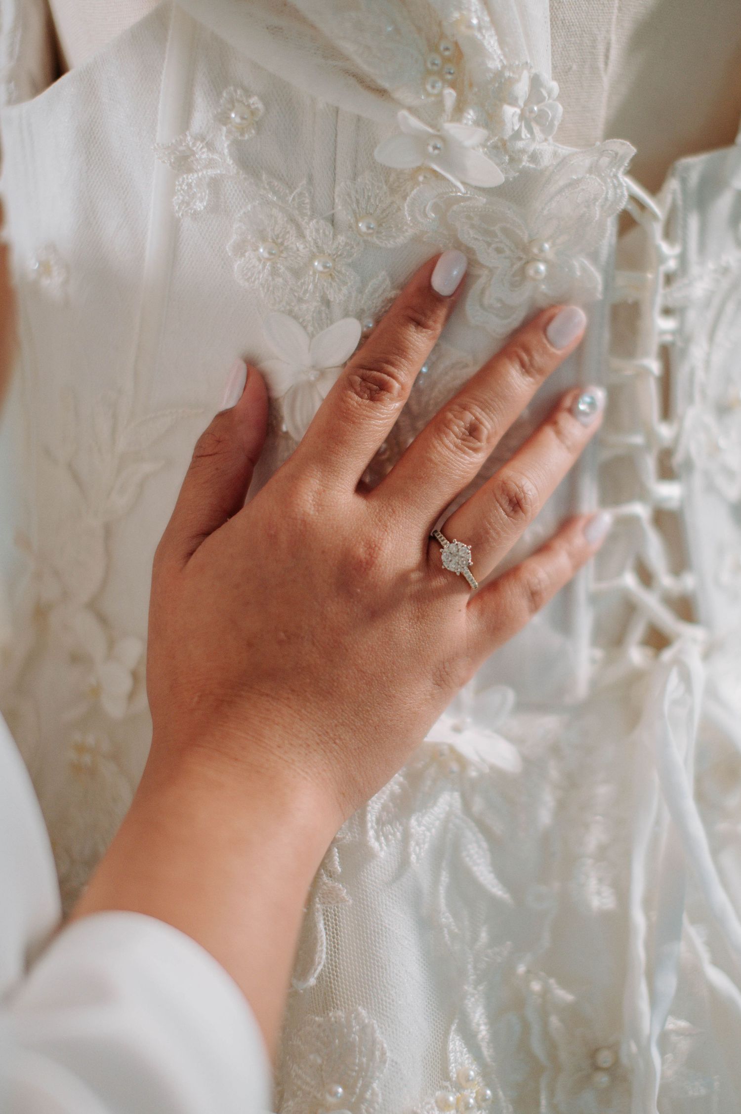 A hand wearing an engagement ring touches delicate white lace details on a wedding dress.