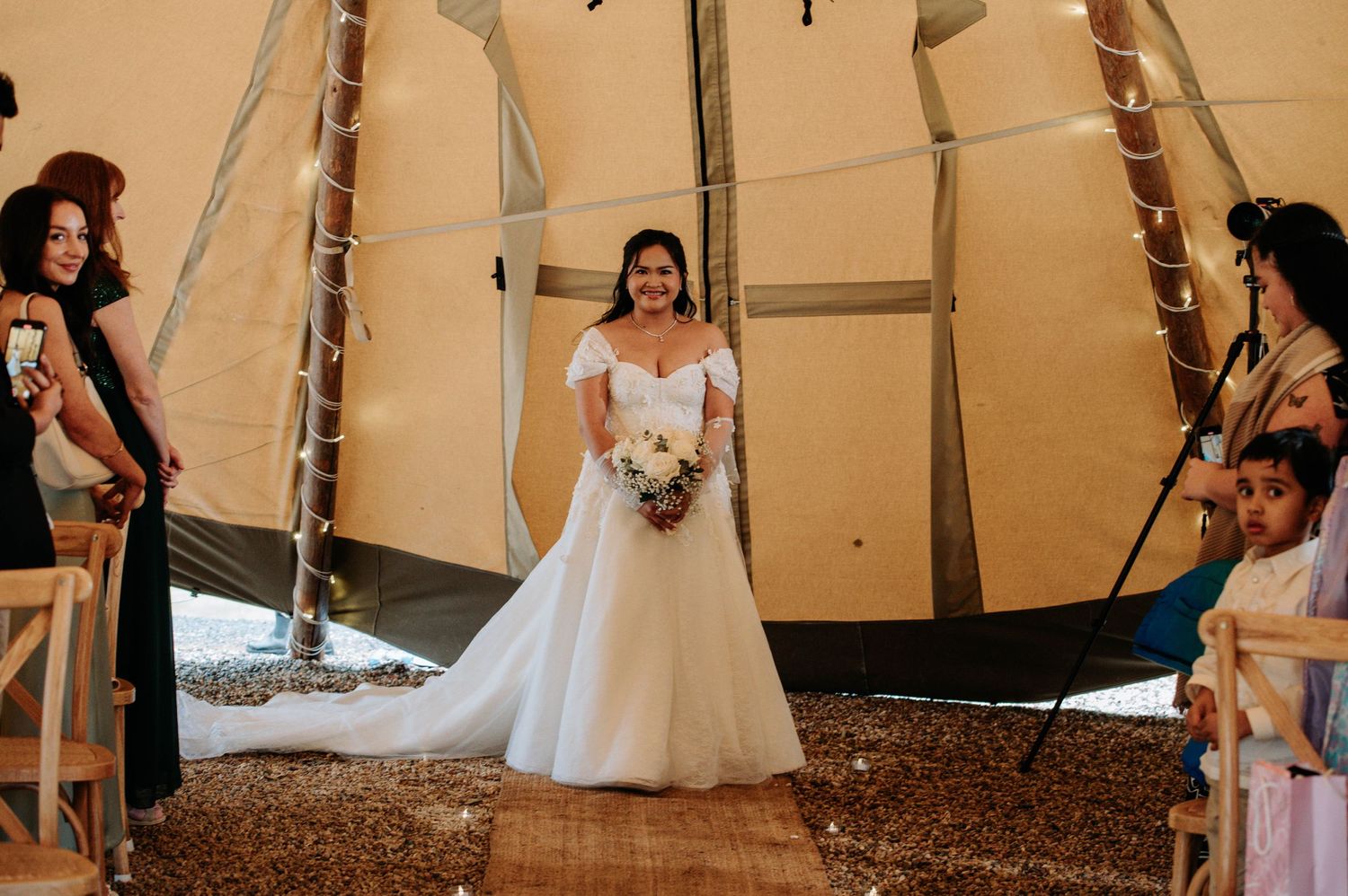 A bride stands in a beautiful white gown inside a decorated teepee during her wedding ceremony.
