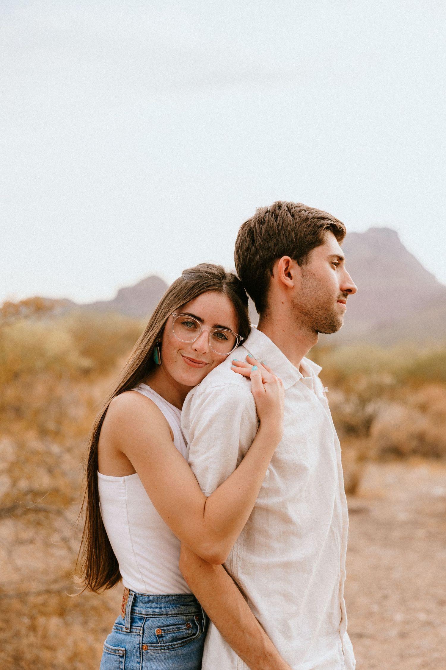 Couple embraces in casual attire against desert mountain backdrop during sunset.