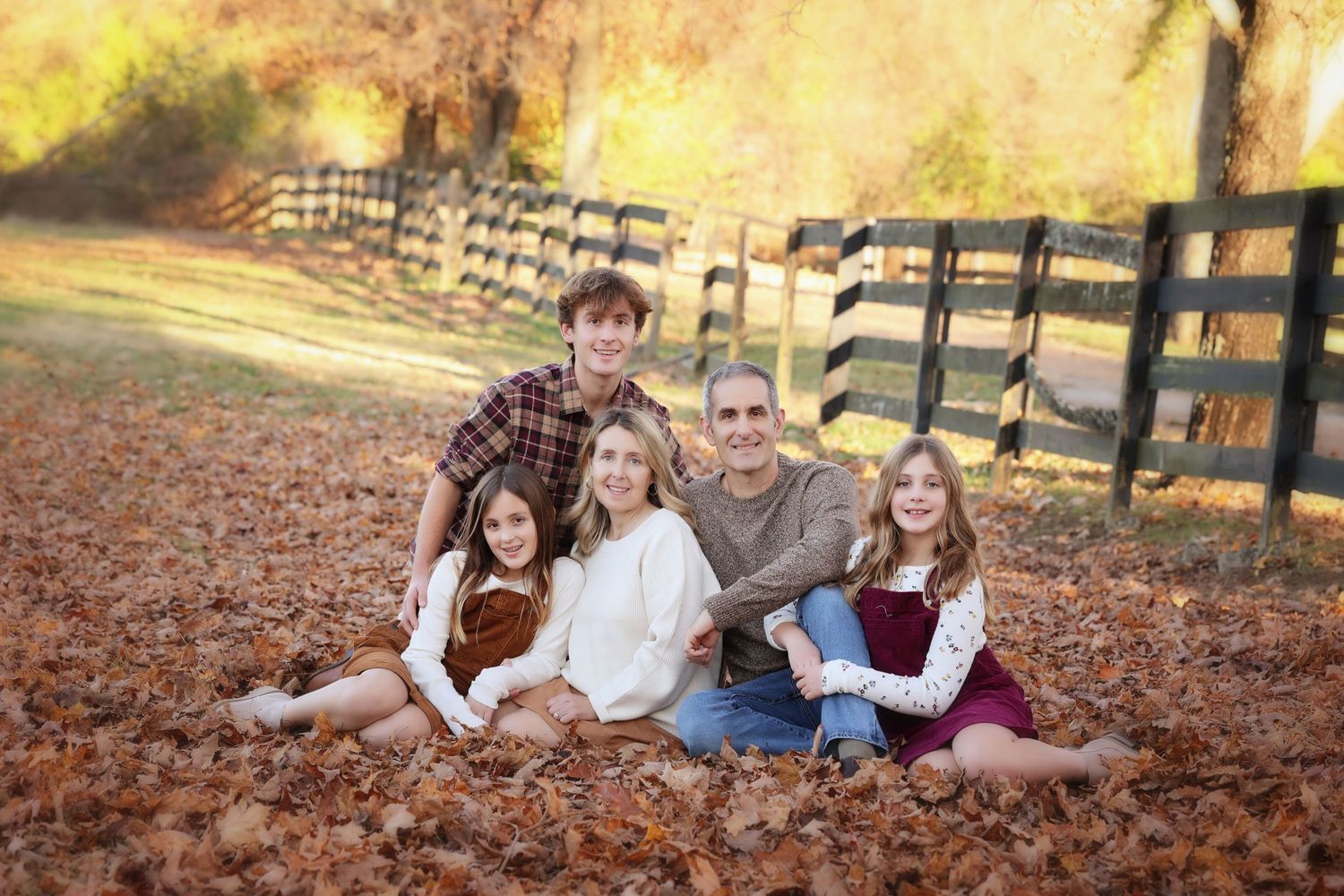 Family group sitting together on fallen leaves with a wooden fence in the background in Custom Fall Family session.