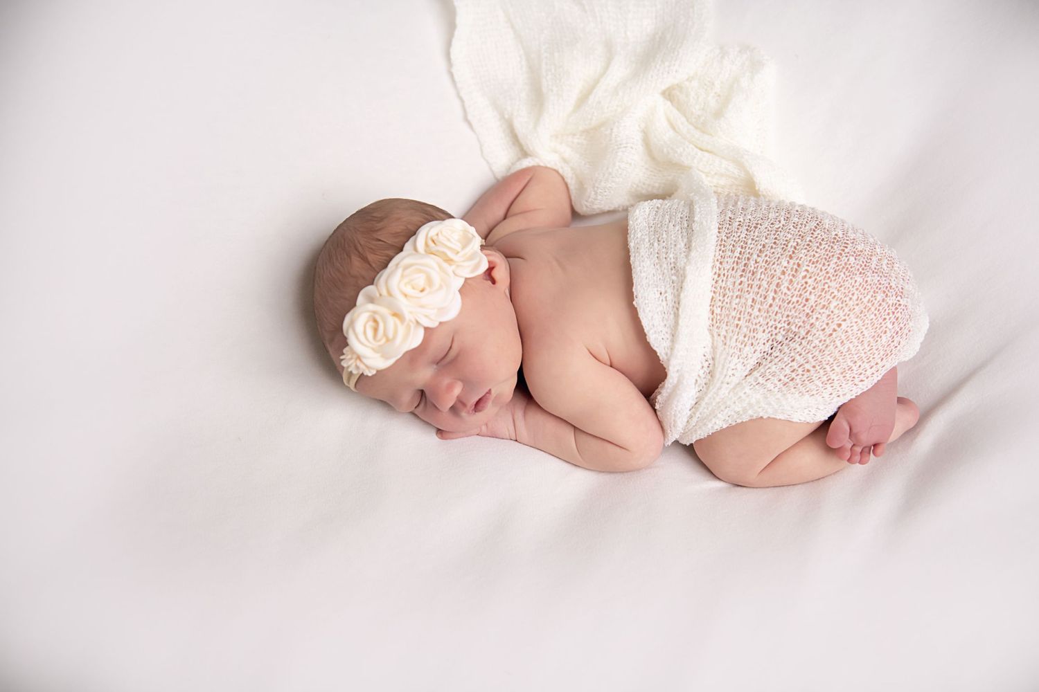 Sleeping newborn baby wrapped in white lace wearing a delicate floral headband.