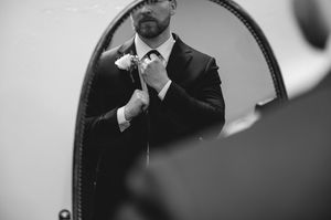 Groom in black suit adjusting tie reflected in round mirror, black and white wedding photo.