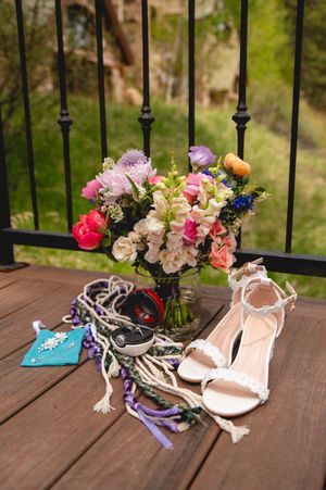 A colorful floral bouquet in glass vase with white sandals and necklaces displayed on wooden deck with metal railings.