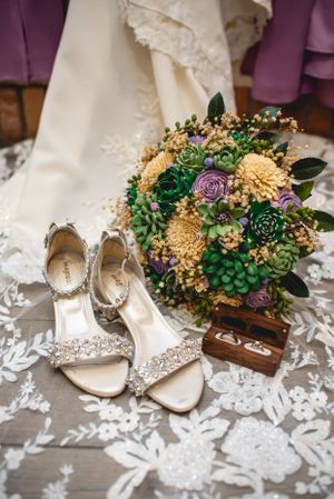 Wedding bouquet with succulents and purple flowers next to sparkly bridal shoes on lace fabric.