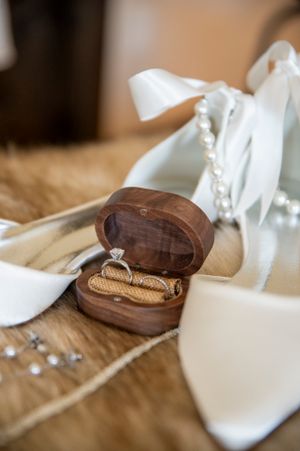 Wedding accessories including white bridal shoes, a wooden ring box with diamond ring, and delicate white ribbons.