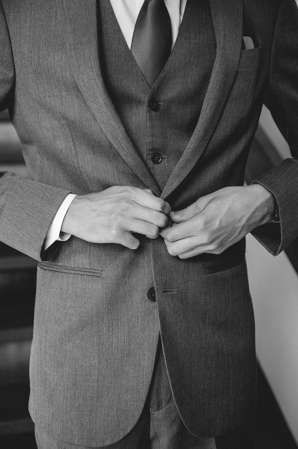 Man in gray three-piece suit buttoning up his formal jacket, shown in elegant black and white photography.