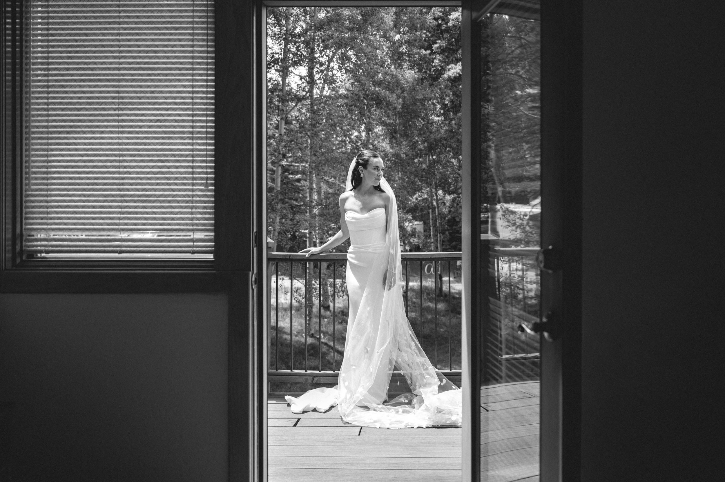 Black and white photo of a bride in a white gown posing on a wooden balcony surrounded by trees.