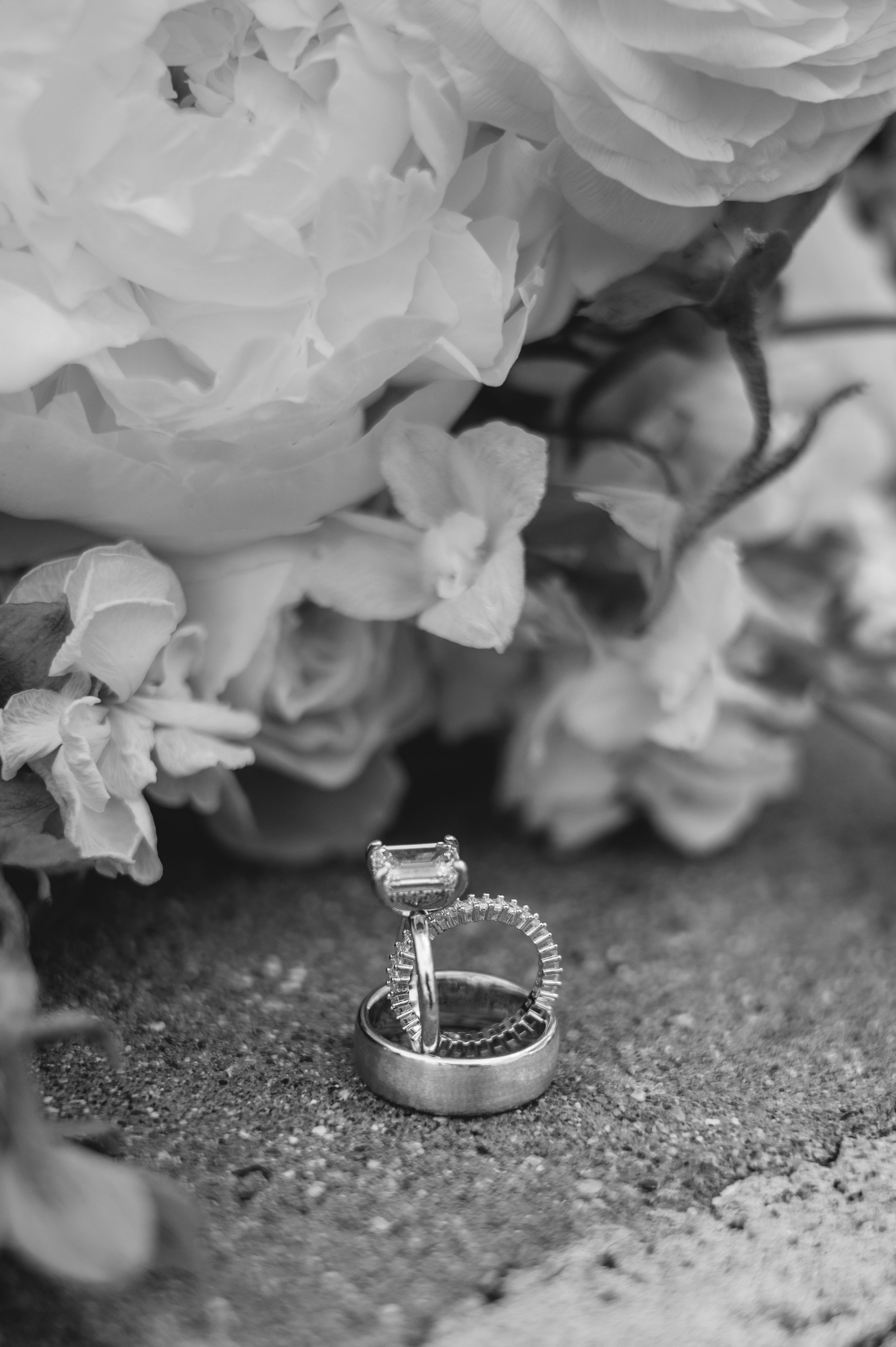 Black and white photo of wedding rings stacked together with a floral bouquet of white roses in the background.