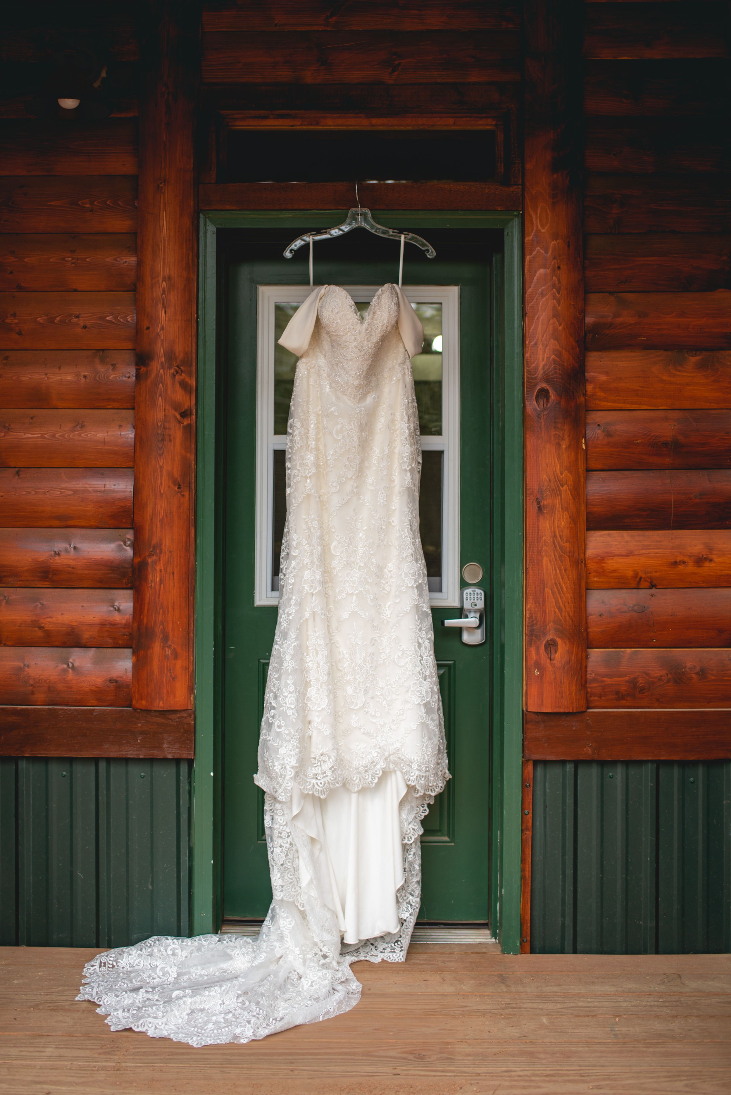 White lace wedding dress hanging on rustic log cabin door with green trim.