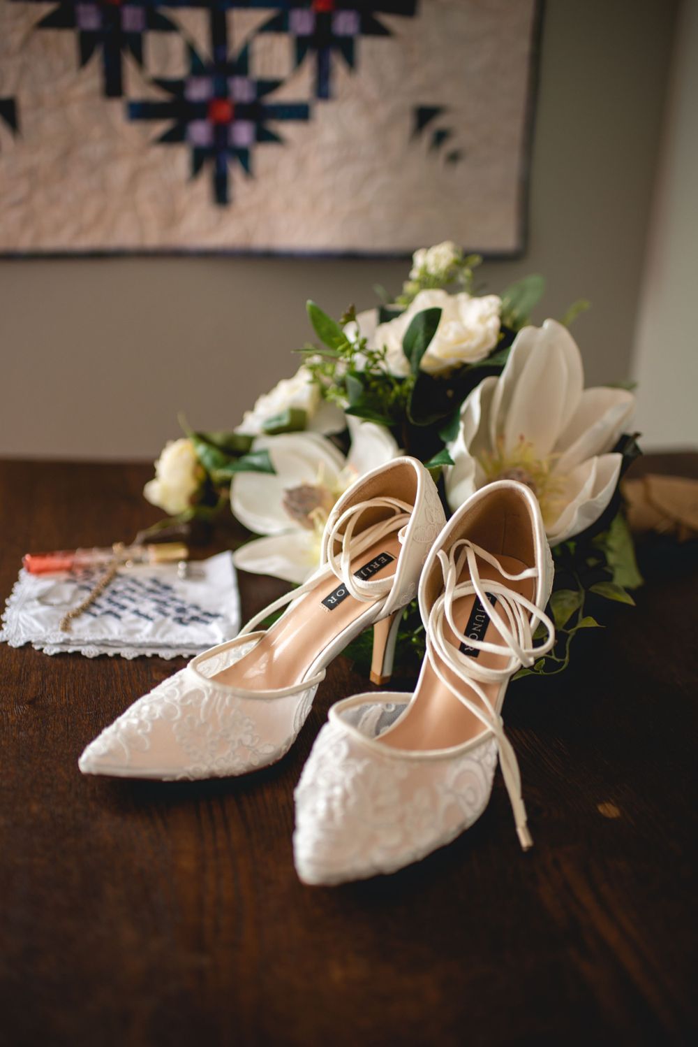 White lace bridal shoes with floral arrangements on dark wooden table.