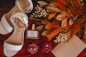 White wedding shoes and accessories arranged with orange lilies and decor on dark wood surface.