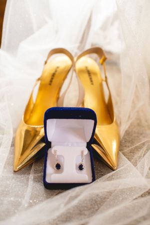 Gold pointed-toe heels with navy jewelry box containing pearl earrings on white tulle fabric.