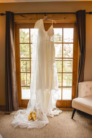 White lace wedding dress hanging in window with golden shoes beneath.