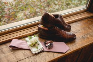 Brown leather dress shoes with wedding rings and pink accessories on wooden windowsill.
