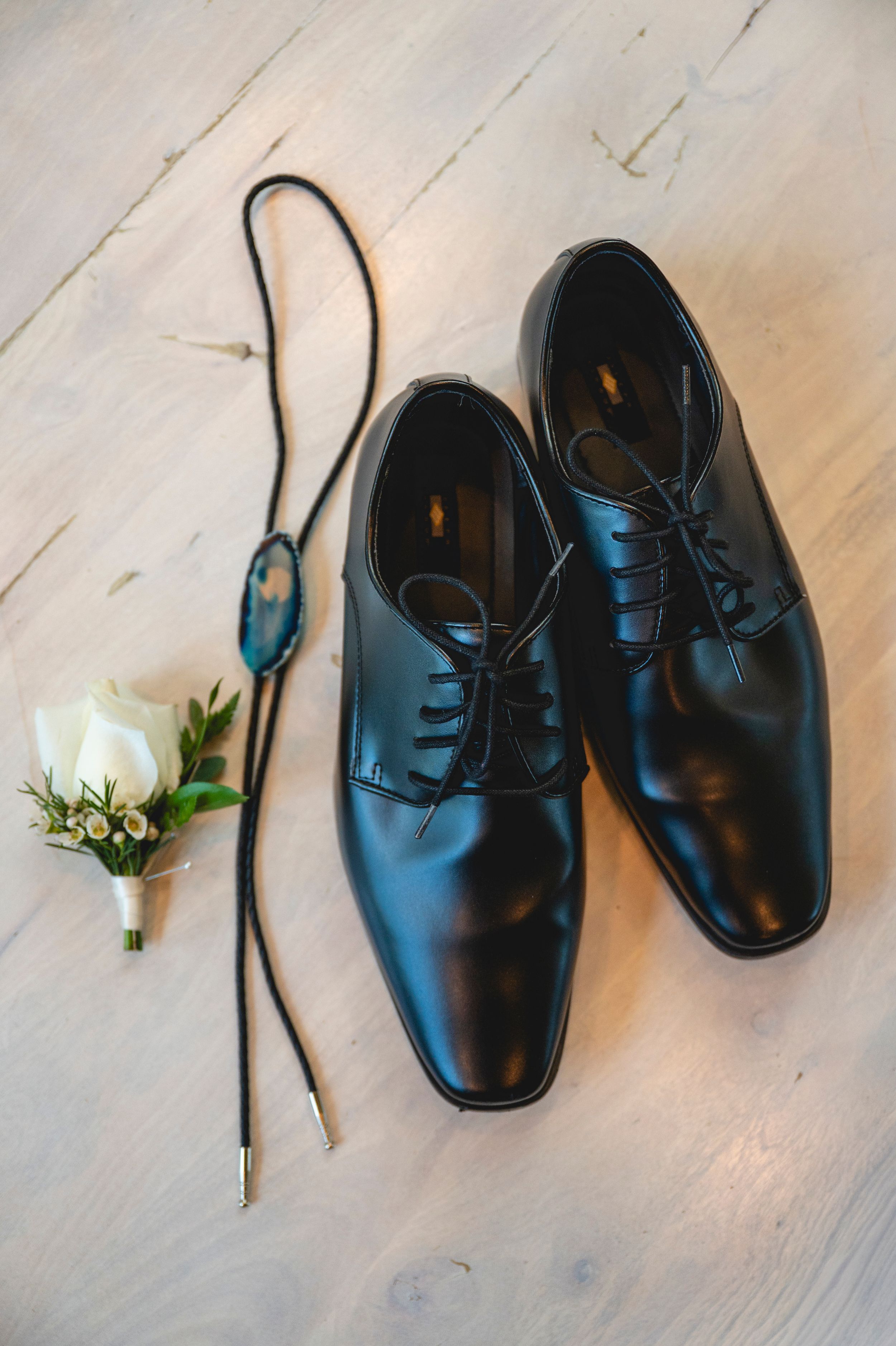 Black leather oxford dress shoes displayed with a bolo tie and white rose boutonniere on a light wood floor.
