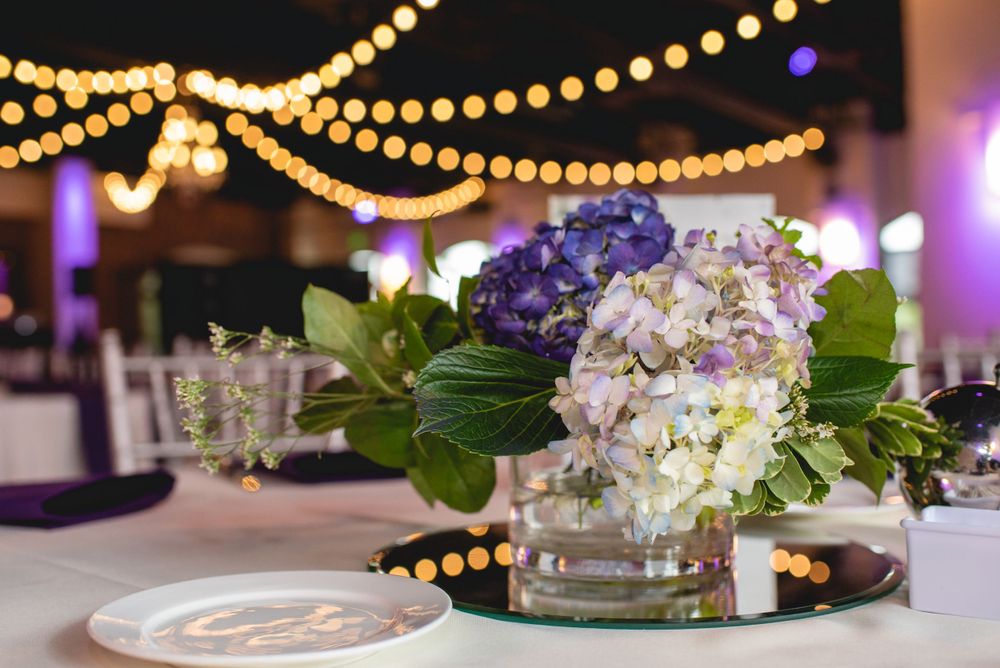 Elegant wedding reception table decor featuring white and purple hydrangeas with string lights overhead.