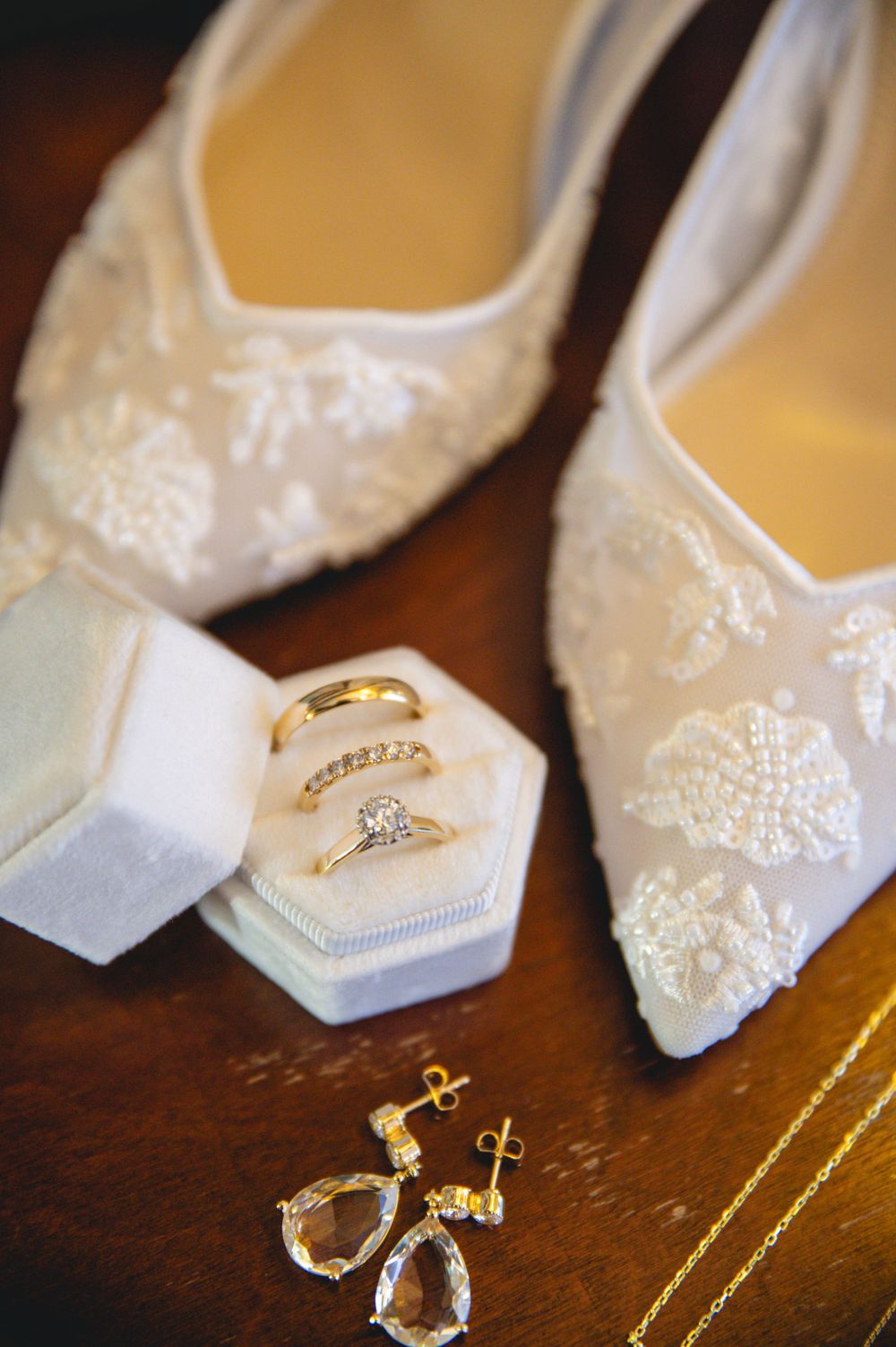 Delicate lace wedding shoes with jewelry and rings displayed on a wooden surface.
