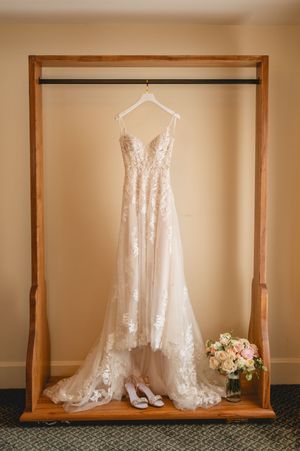 Elegant white lace wedding dress hangs on wooden frame with white heels and blush floral bouquet beneath it.