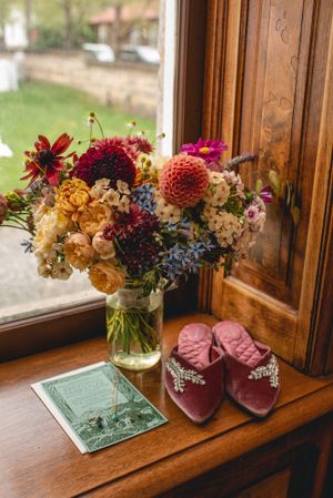 Colorful wildflower bouquet in glass vase with pink slippers on wooden windowsill.