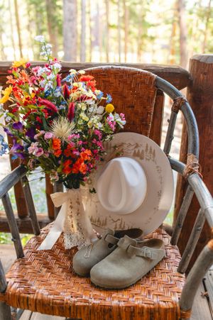 A vibrant wildflower bouquet sits on a rustic wicker chair with a white sunhat and gray shoes in natural light.