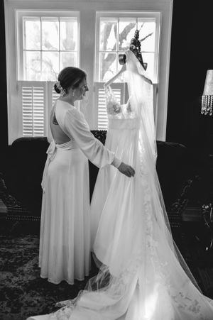 A white wedding gown hangs elegantly by a bright window as preparations take place.