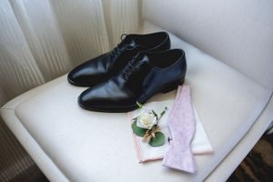 Black leather oxford shoes with pink napkin and floral boutonniere on white chair.