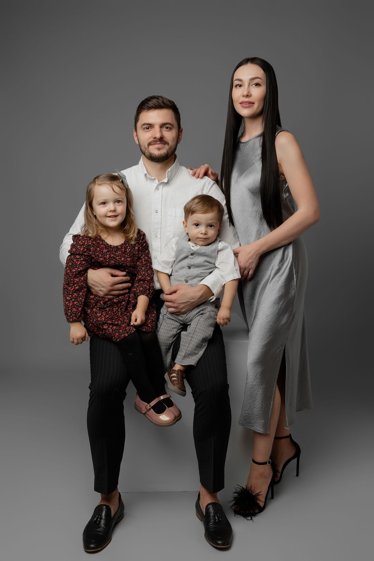 Modern family portrait with parents and two young children in coordinated outfits against grey backdrop.