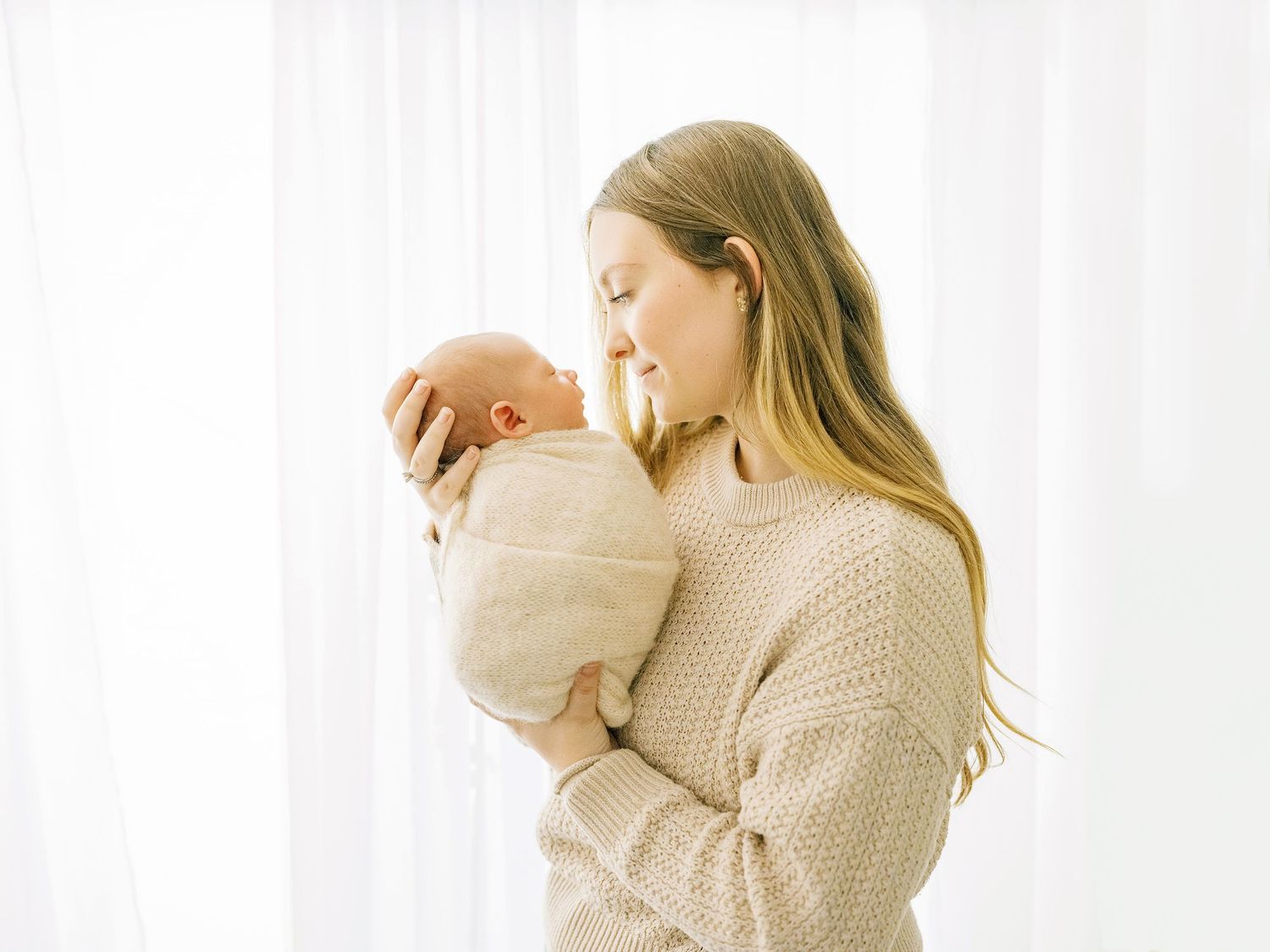 A person in a cream sweater holds and tenderly embraces a newborn baby against a bright white background.