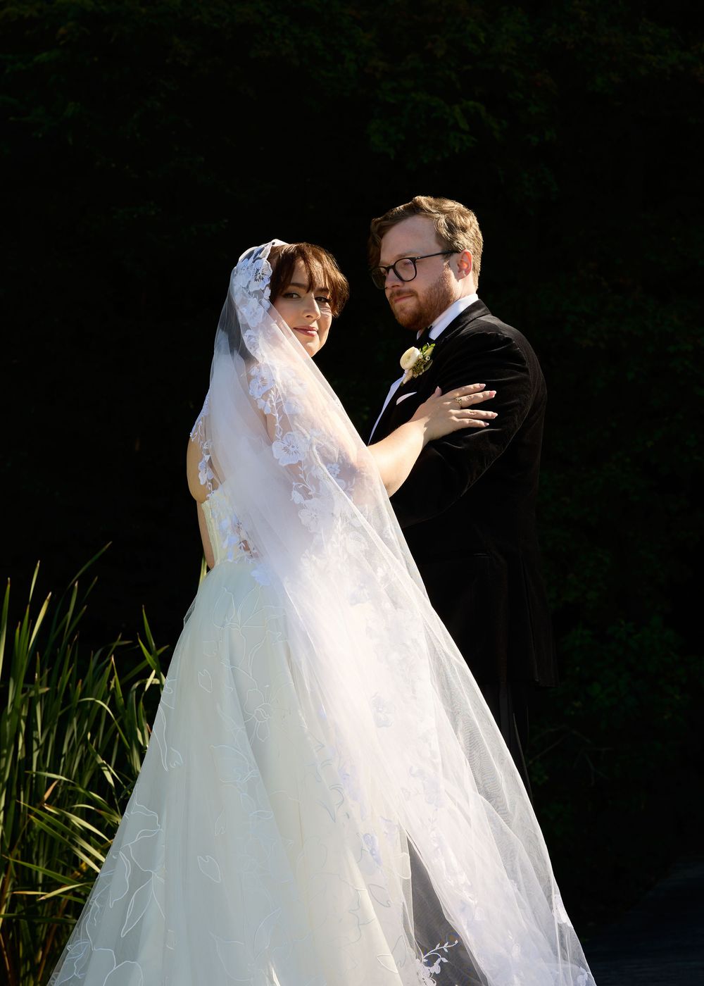 A bride and groom share an intimate moment outdoors in dramatic lighting with a long flowing white veil and dark background.