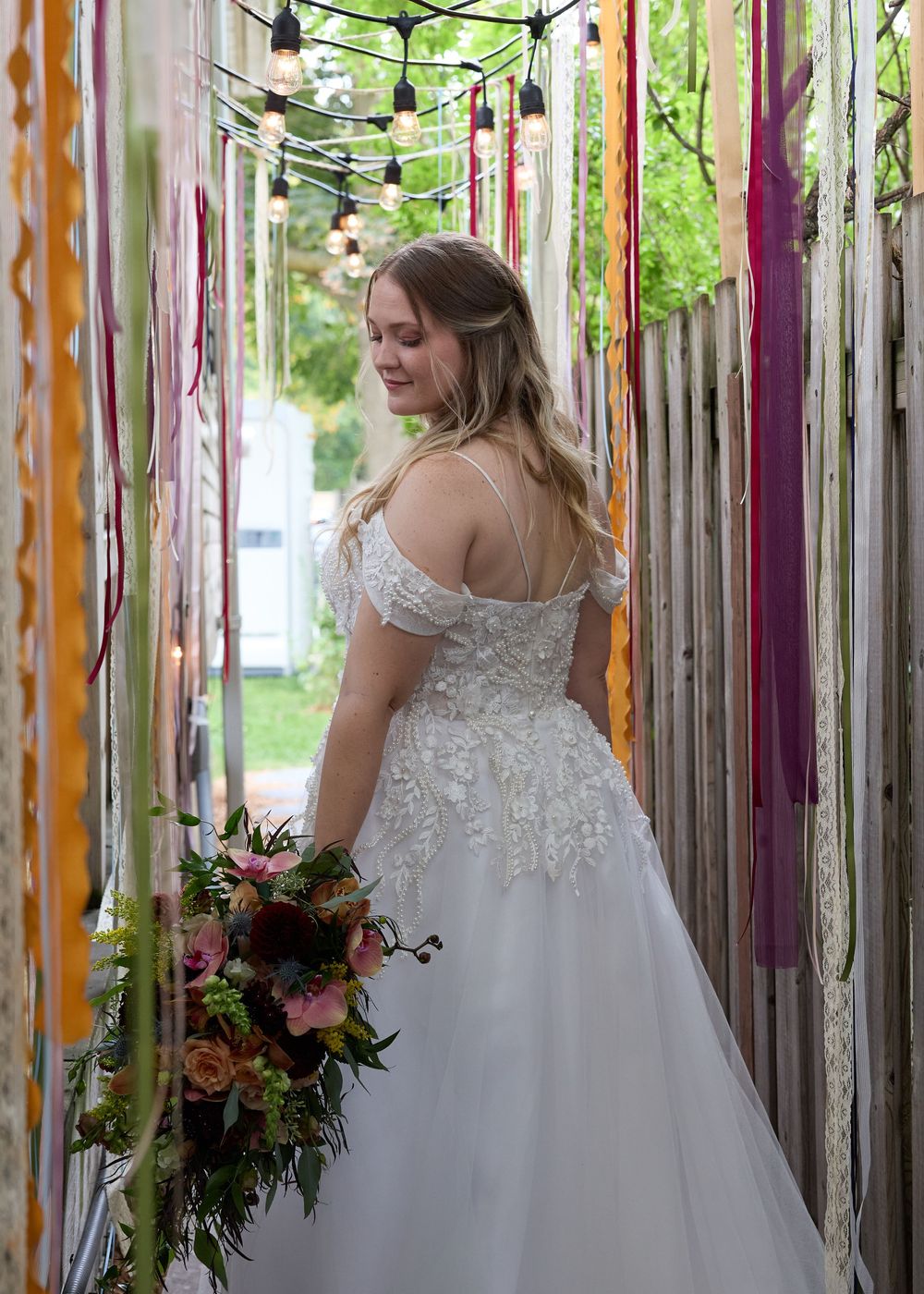 A person in a white lace wedding dress stands in a whimsical outdoor space with colorful ribbons and hanging lights.