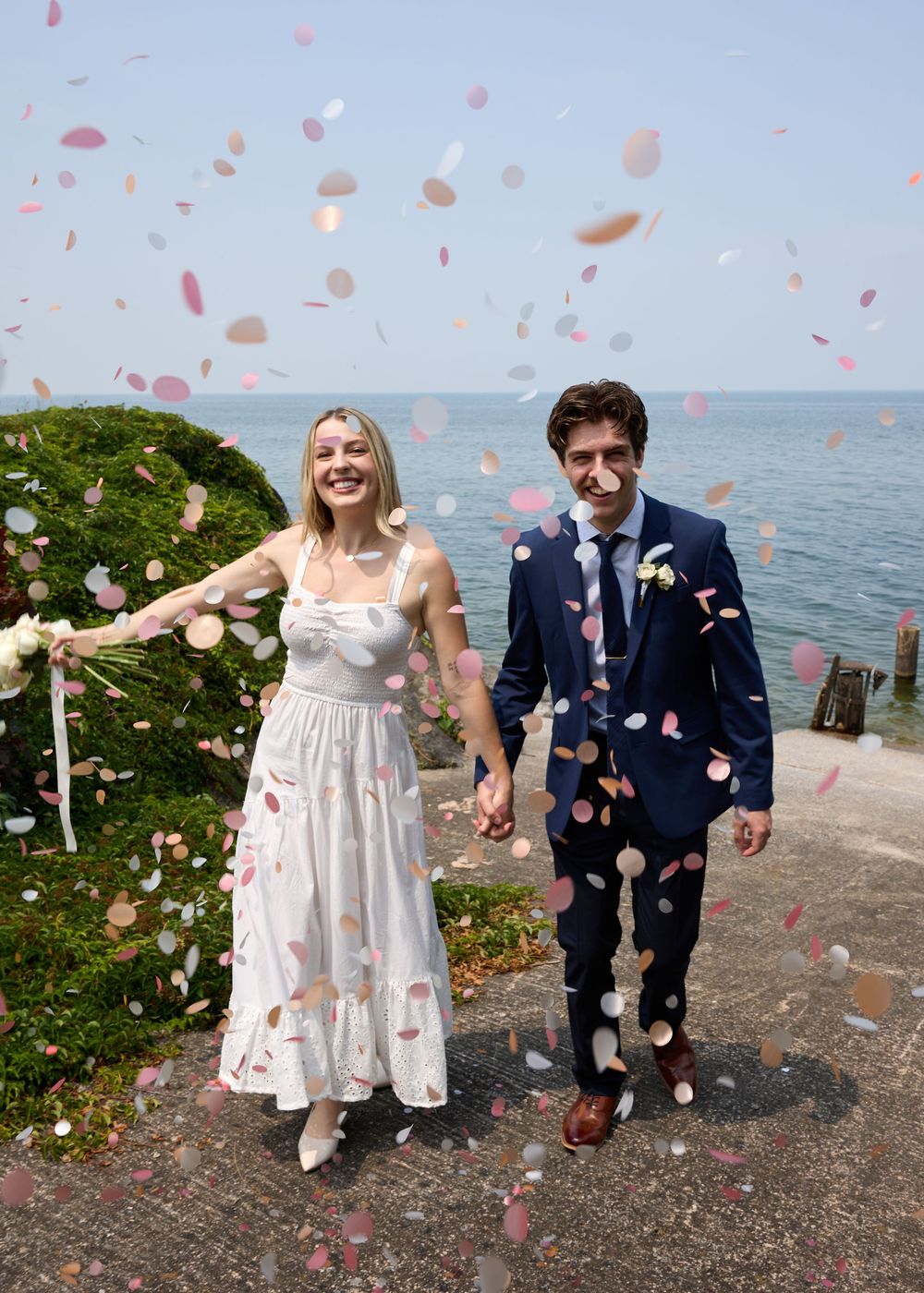 A couple walks along a waterfront path while pink flower petals fall around them in a romantic celebration.