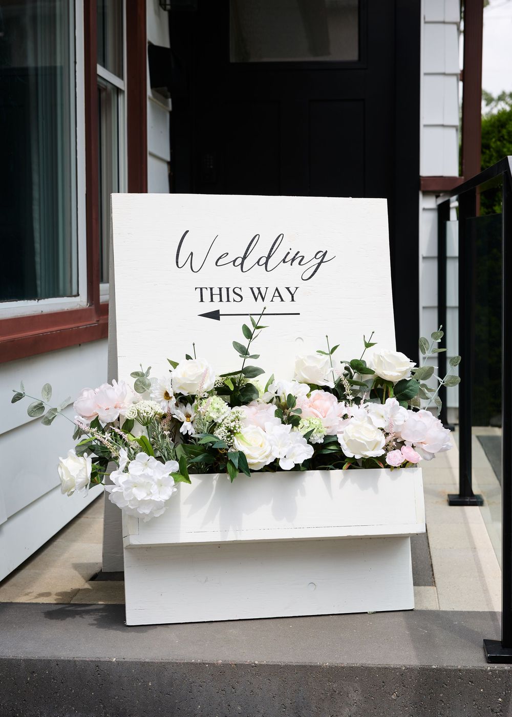 White wedding directional sign adorned with pink and white roses in a floral box arrangement on a porch entrance.