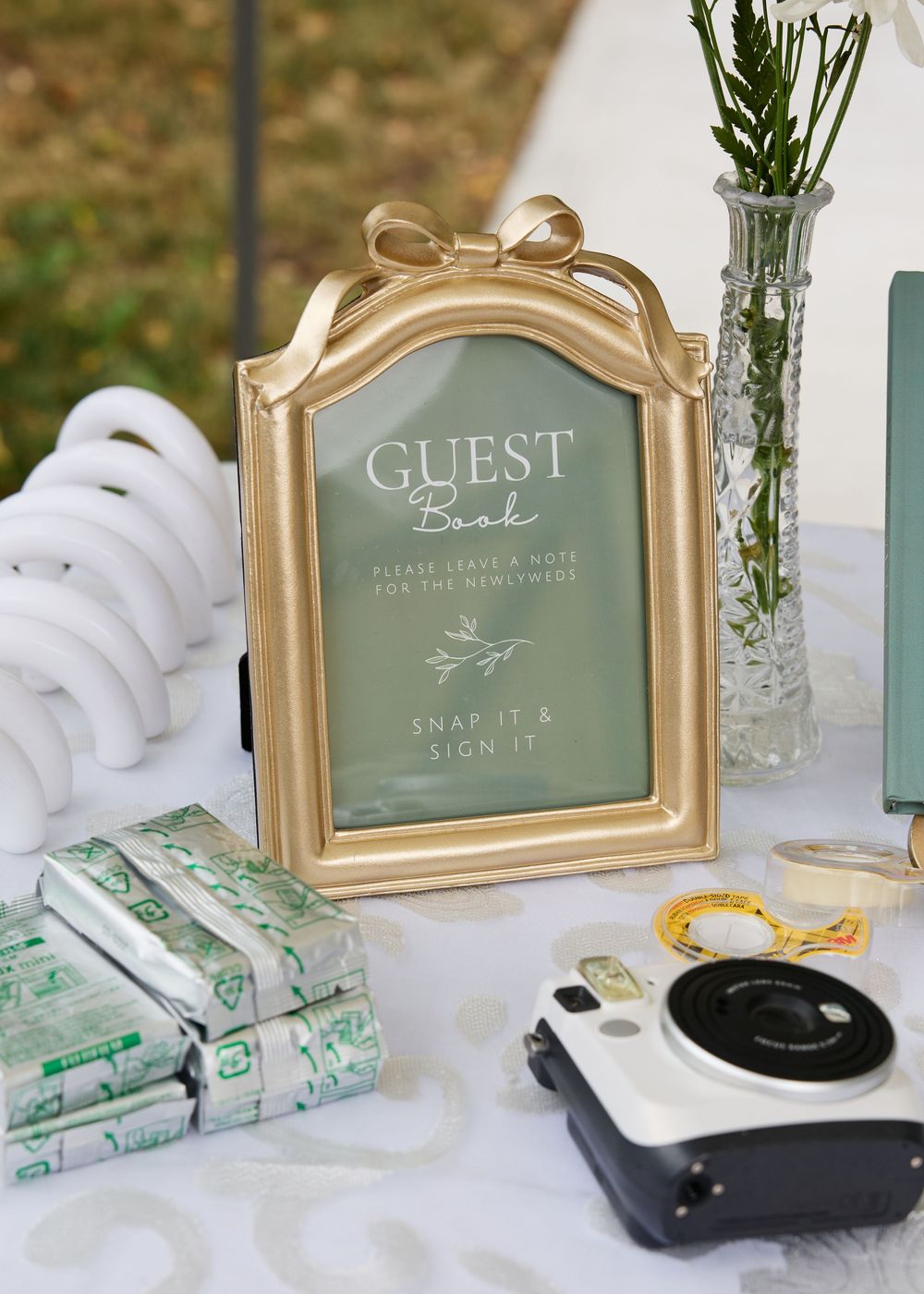 Gold framed guest book sign with Instax camera and film rolls on wedding reception table with greenery.