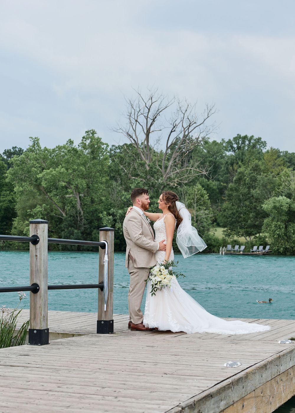 Bride and groom share a romantic moment on a wooden dock overlooking a turquoise lake surrounded by lush green trees.
