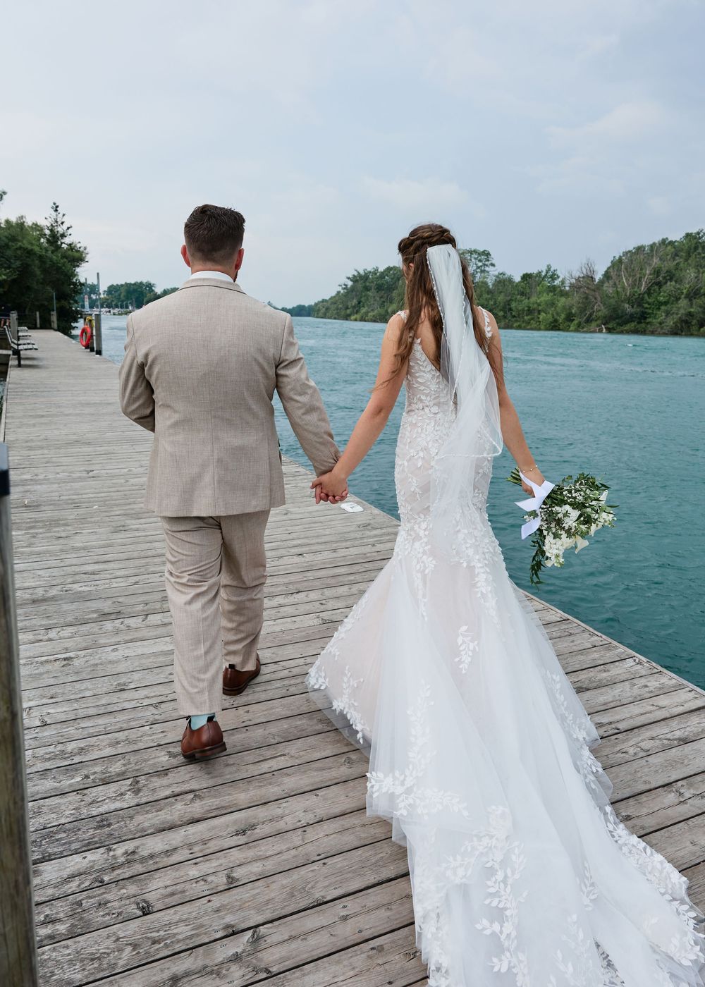 Bride and groom walking hand-in-hand on a wooden dock by turquoise water, bride in lace mermaid gown with veil.