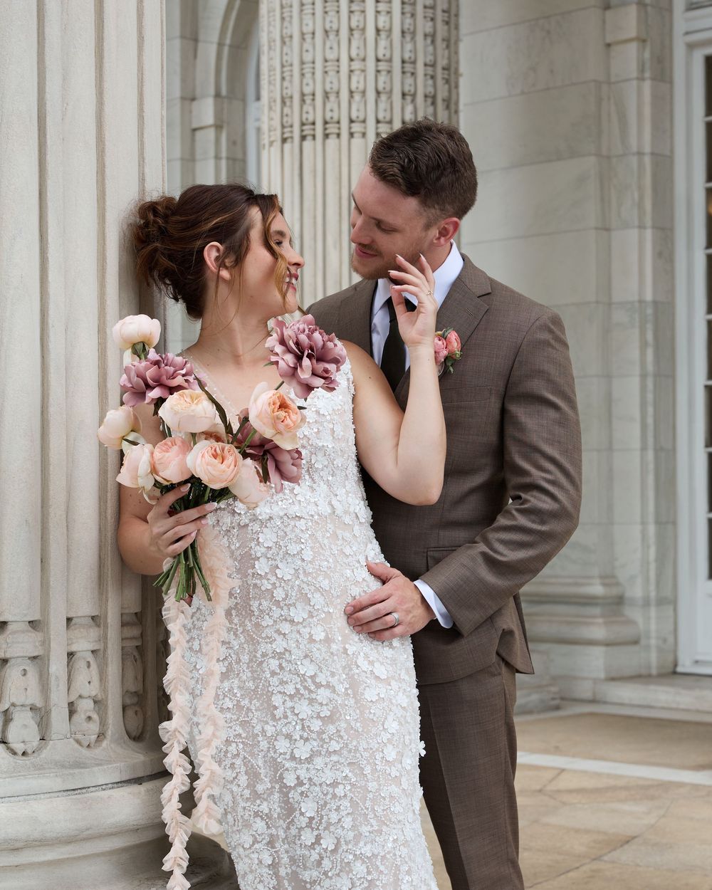 Romantic newlywed couple sharing an intimate moment at a stunning marble stone building with floral bouquet decorations.