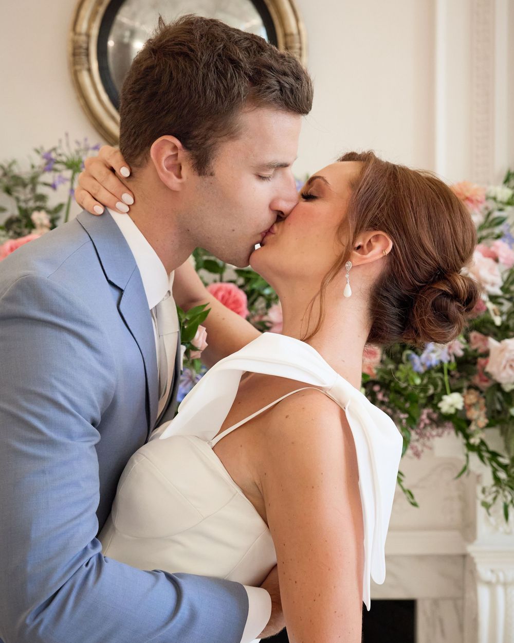 A couple in formal attire share a romantic kiss at their wedding ceremony with flowers and decorations in the background.