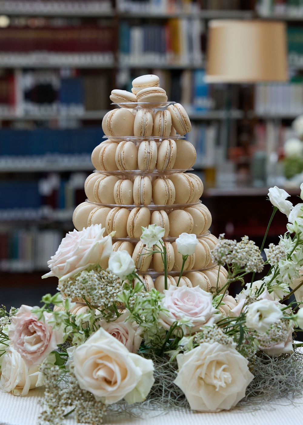 A tower of beige macarons decorated with delicate roses and baby's breath flowers on a blurred library background.