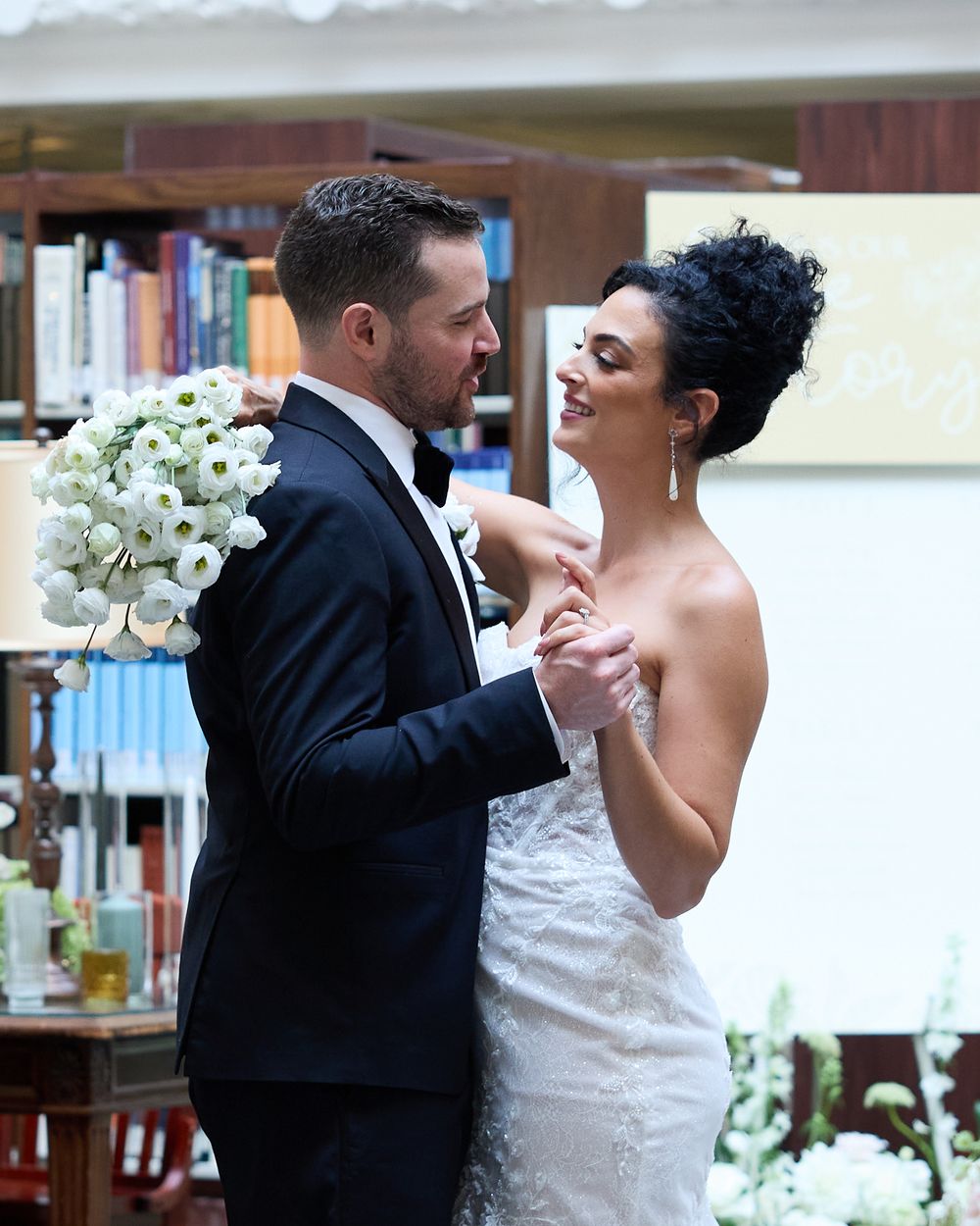 A romantic wedding dance moment showing a bride in white lace and groom in black suit with white flowers in the background.